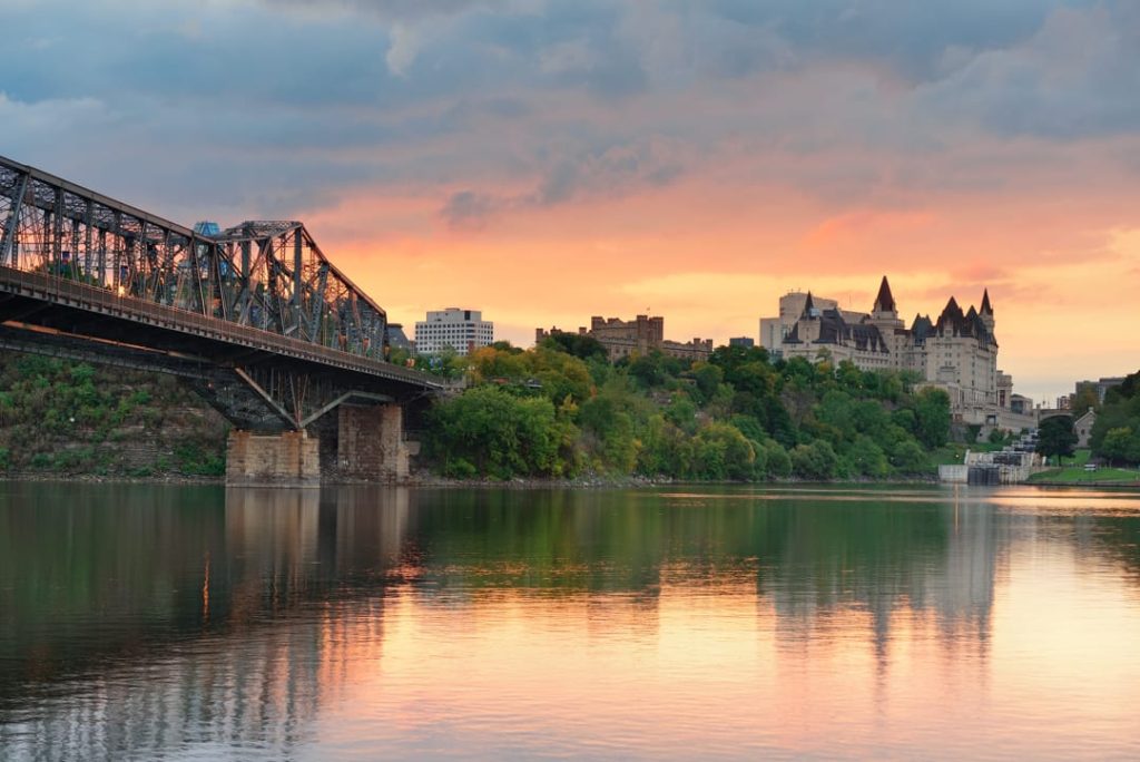 IJzeren spoorbrug over kalm water met de skyline en Château Laurier van Ottawa op de achtergrond bij zonsondergang