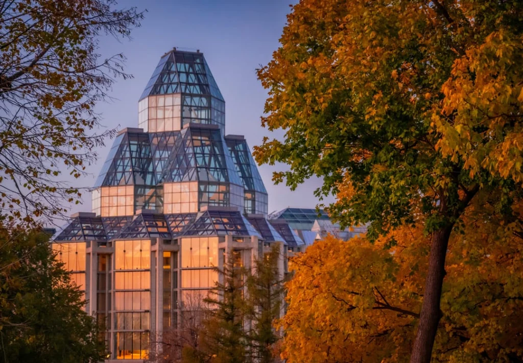 Glazen torenspits van het kunstmuseum National Gallery of Canada, gefotografeerd tussen bomen in oranje herfstkleuren bij avondlicht