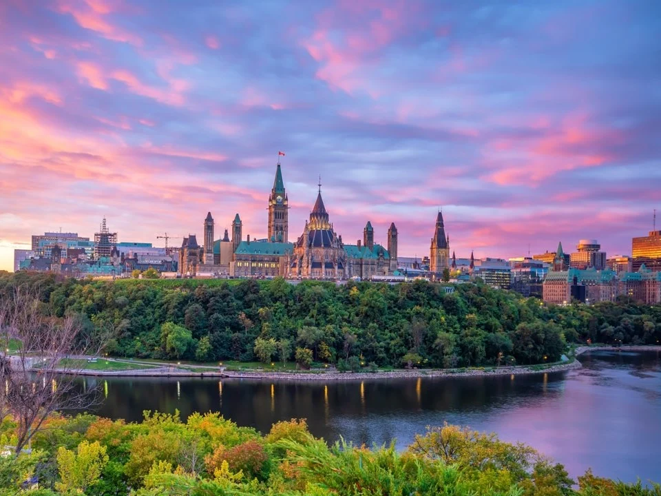 Silhouet van het Canadese parlementsgebouw in Ottawa tegen een roze en paarse avondlucht met de rivier op de achtergrond
