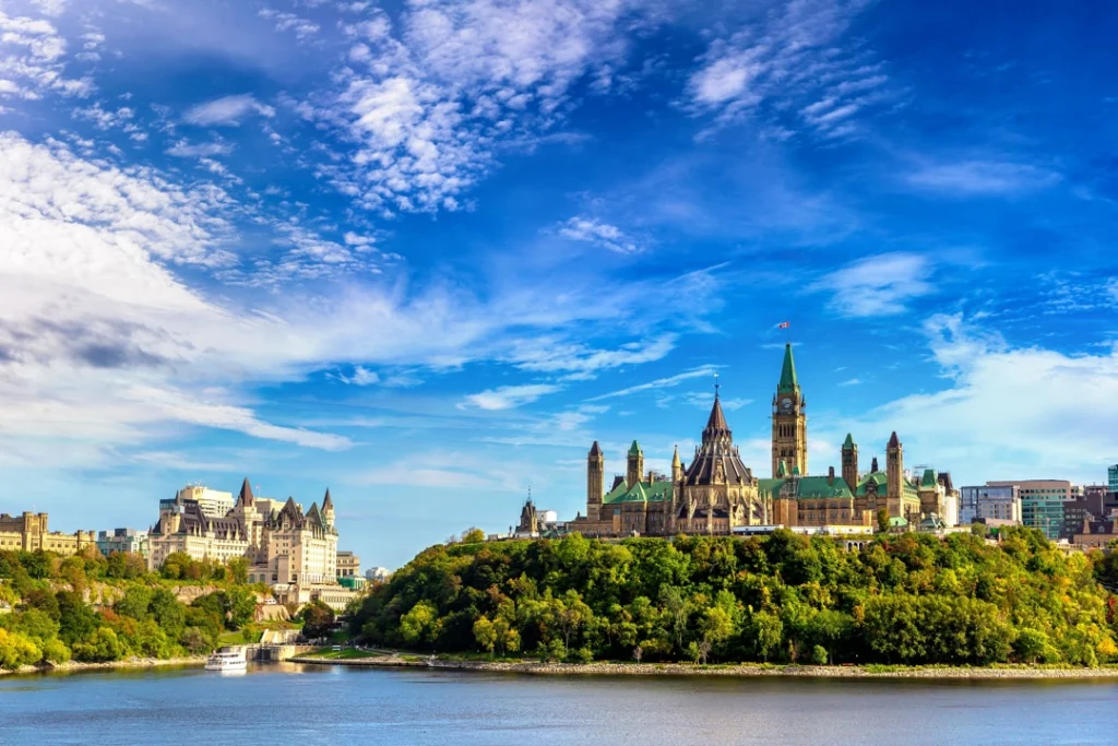 Parlementsgebouwen van Ottawa op een groene heuvel aan de rivier, met een boot op het water en wolken