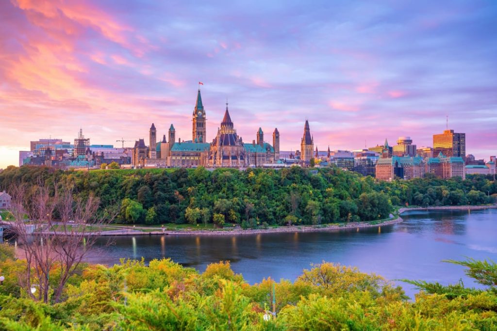 De gebouwen van Parliament Hill met koperen daken op een heuvel boven de rivier, gefotografeerd bij paarsroze schemerlicht in Ottawa
