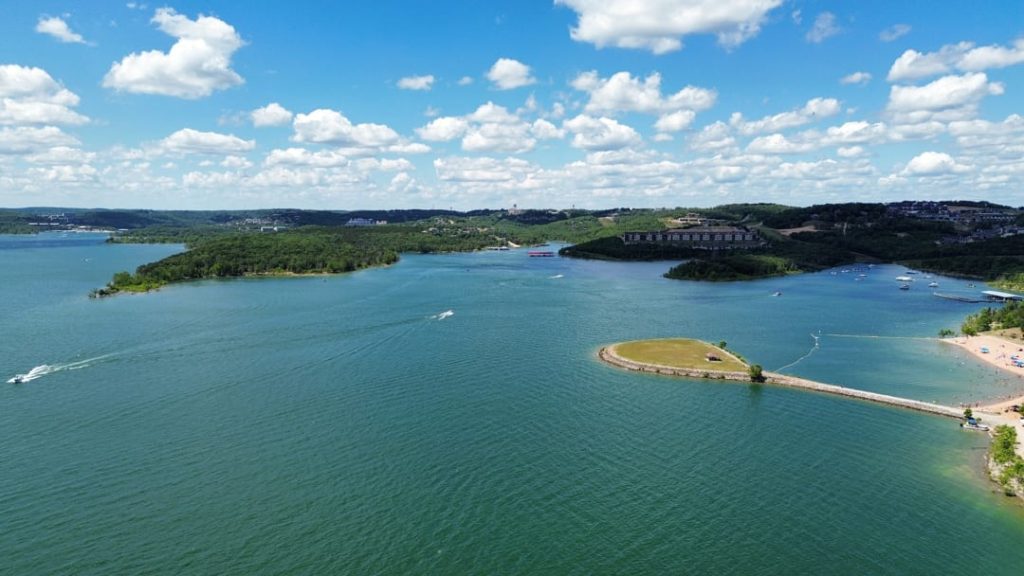 Turkooisblauw meer met een rond schiereiland, vaarende bootjes en beboste oevers met vakantiehuizen in de verte