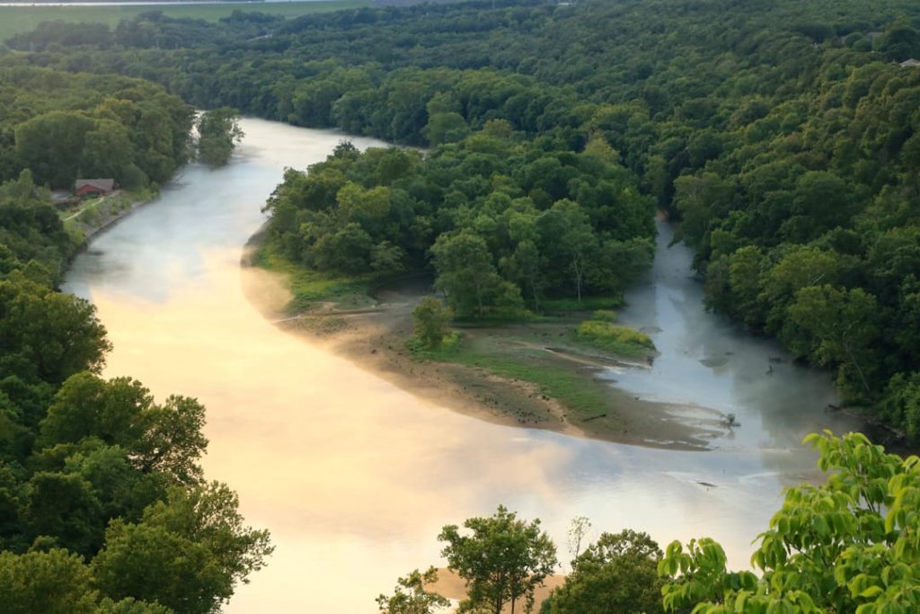 Rivier met lichte nevel boven het water, omringd door groene beboste heuvels en een zandbank met een boomeilandje in de Ozarks
