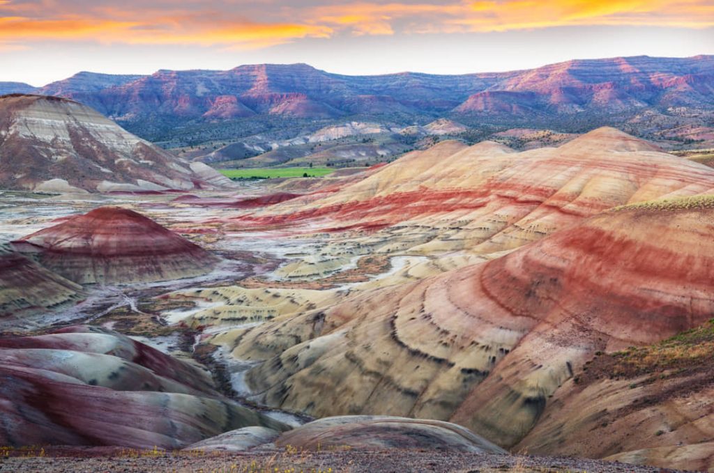 Gelaagde rode en gele heuvels van Painted Hills onder een gloeiende oranje avondlucht met bergen op de achtergrond