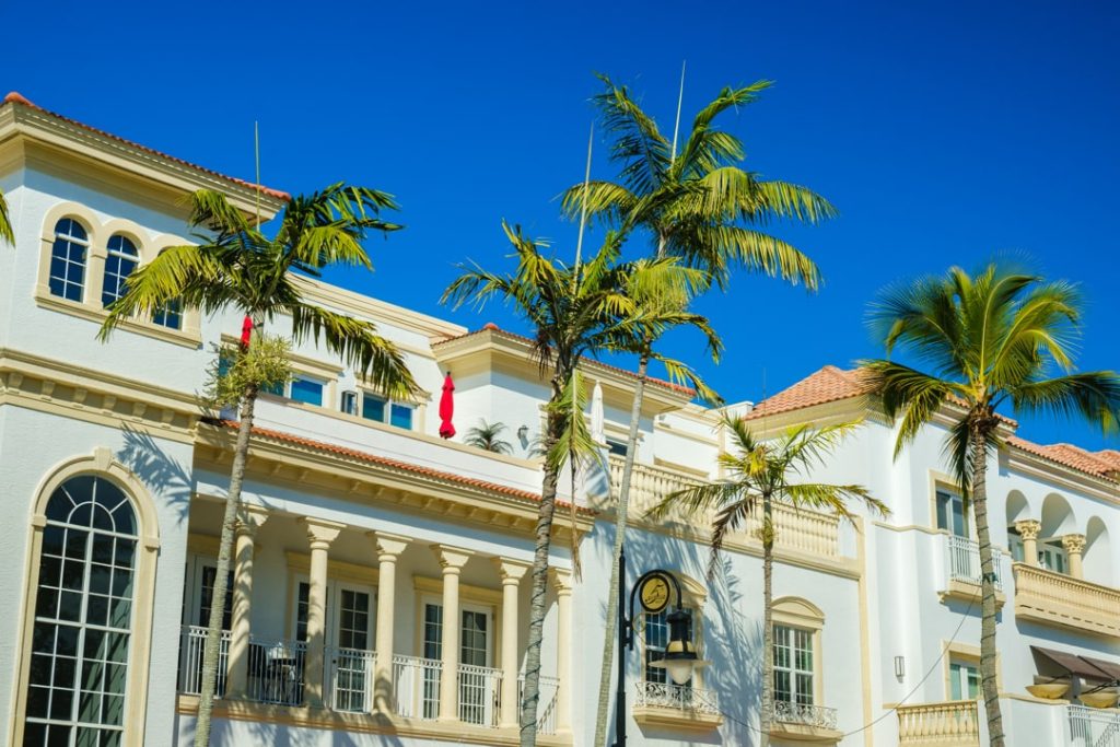 Statig wit gebouw met zuilen en balkons, gezien tussen hoge palmbomen tegen een strakblauwe lucht in Palm Beach