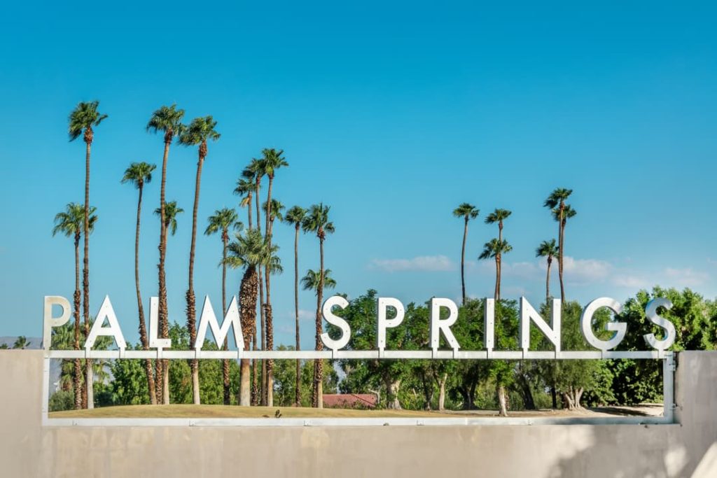 Groot wit bord met de tekst Palm Springs, omringd door hoge palmbomen tegen een strakblauwe lucht