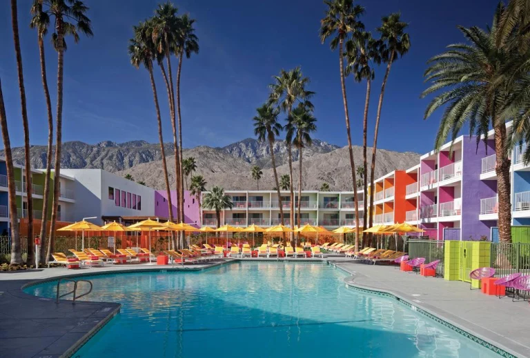 Zwembad omgeven door gele parasols voor felgekleurde hotelgebouwen met hoge palmbomen en bergen op de achtergrond in Palm Springs