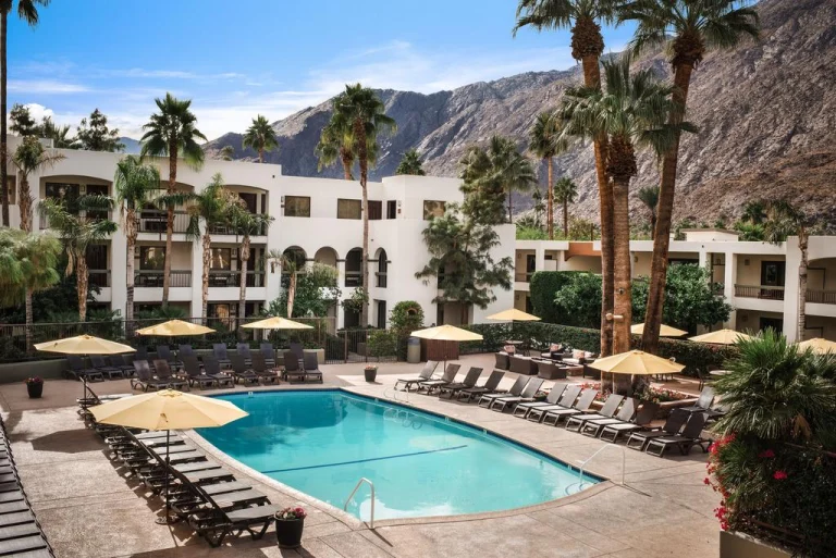 Rond zwembad met gele parasols en ligstoelen bij een wit resort met palmbomen en bergen op de achtergrond in Palm Springs