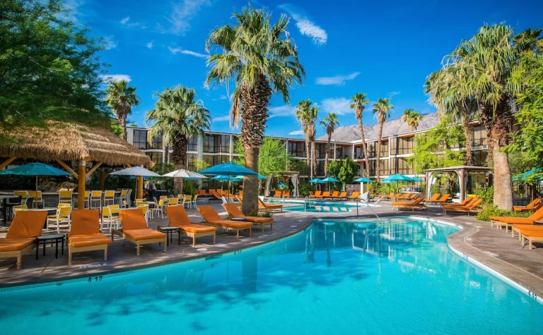 Groot zwembad omringd door ligstoelen, oranje parasols en hoge palmbomen bij een resort in Palm Springs