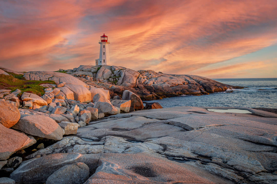 Witte vuurtoren met rode kap staat op gladde granieten rotsen aan de kust van Peggy's Cove terwijl de lucht oranje kleurt bij zonsondergang