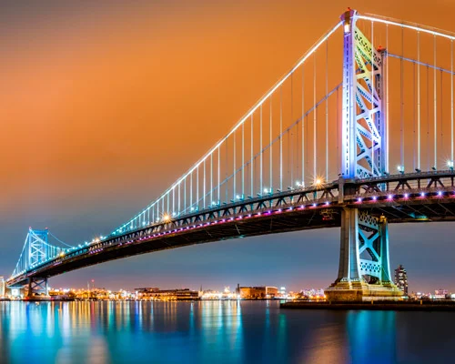 Verlichte hangbrug Benjamin Franklin Bridge over de rivier bij Philadelphia met een oranje avondlucht en weerspiegeling in het water