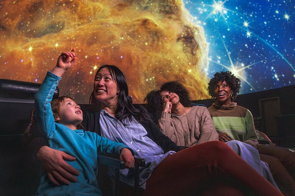 Gezin met een jongen die opgewonden omhoog wijst naar een sterrenhemel op het koepelscherm van het planetarium