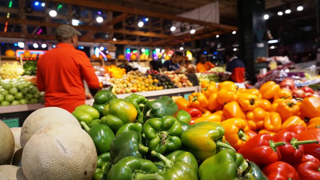 Kraam met stapels groene, gele en rode paprika's en meloenen met bezoekers die tussen de kramen lopen in Reading Terminal Market