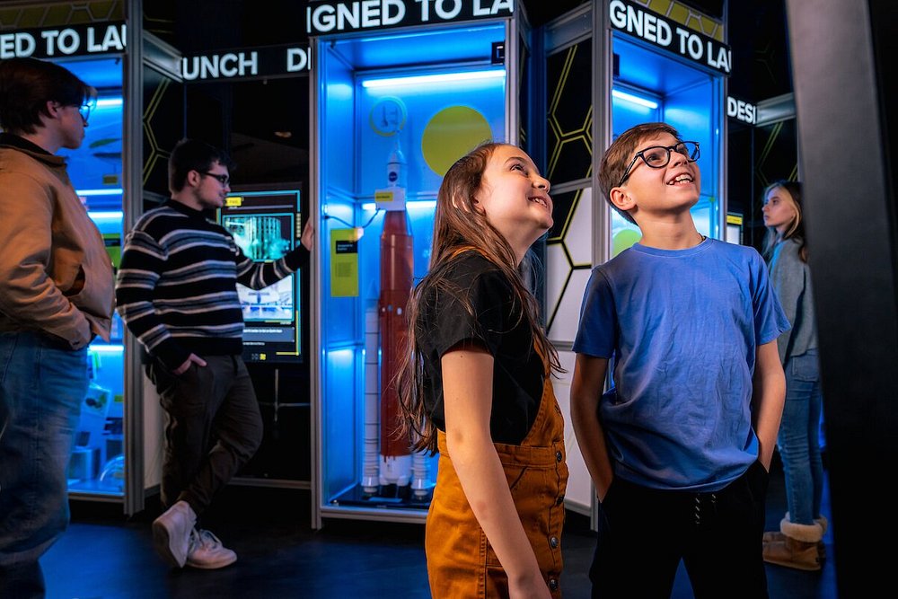 Twee lachende kinderen kijken omhoog naar een verlichte vitrine met een raketmodel in een expositie over ruimtevaart