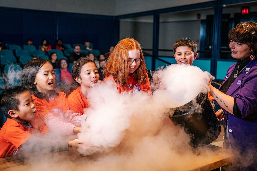 Groep enthousiaste kinderen kijkt naar een wolk witte rook tijdens een sciencedemonstratie met een presentator