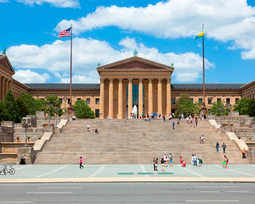 Mensen lopen over de brede stenen trap voor het zuilengebouw van het Philadelphia Museum of Art onder een blauwe lucht met wolken