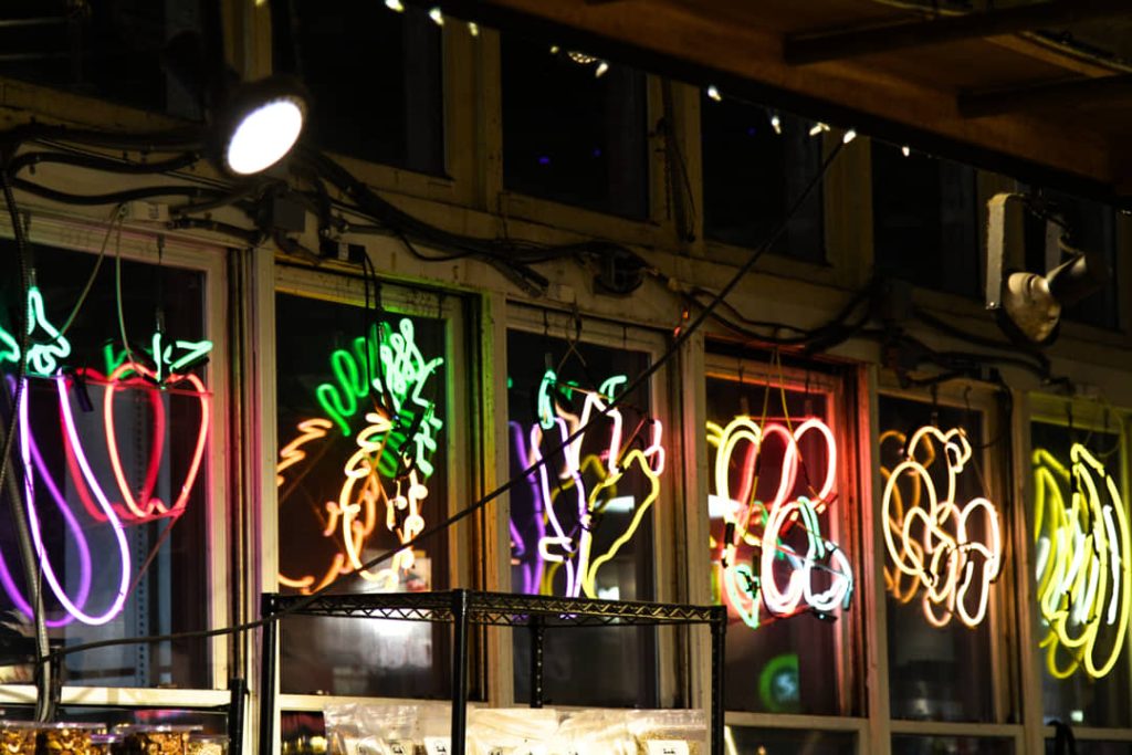 Rij ramen met felgekleurde neonverlichting in de vorm van paprika's, ananassen en ander fruit in Reading Terminal Market bij avond