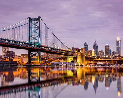 De verlichte hangbrug Benjamin Franklin Bridge met op de achtergrond de skyline van Philadelphia weerspiegeld in het water bij avondschemering