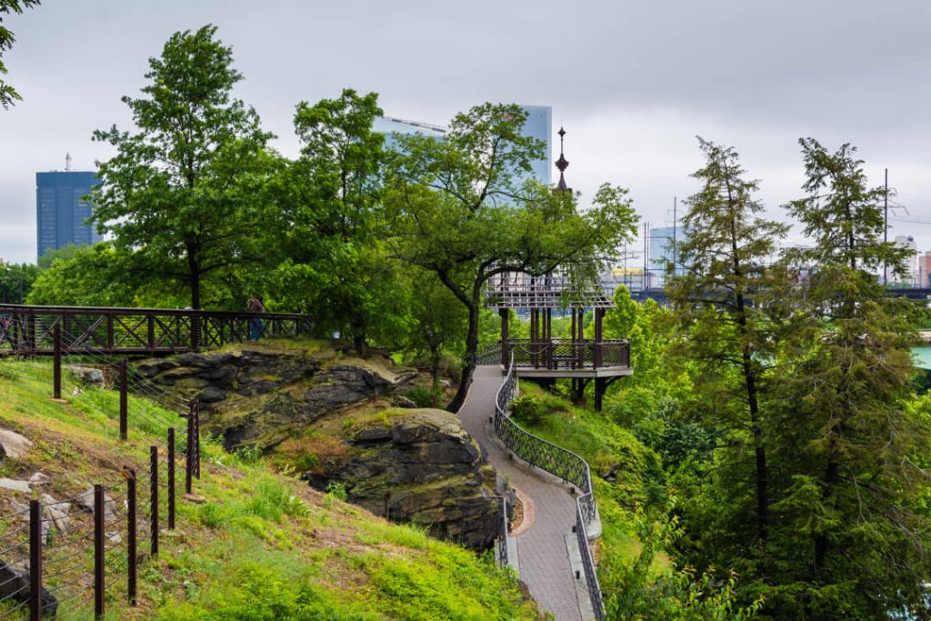 Kronkelend pad met een houten brug over een rotsachtige heuvel in een groen park met wolkenkrabbers in de verte