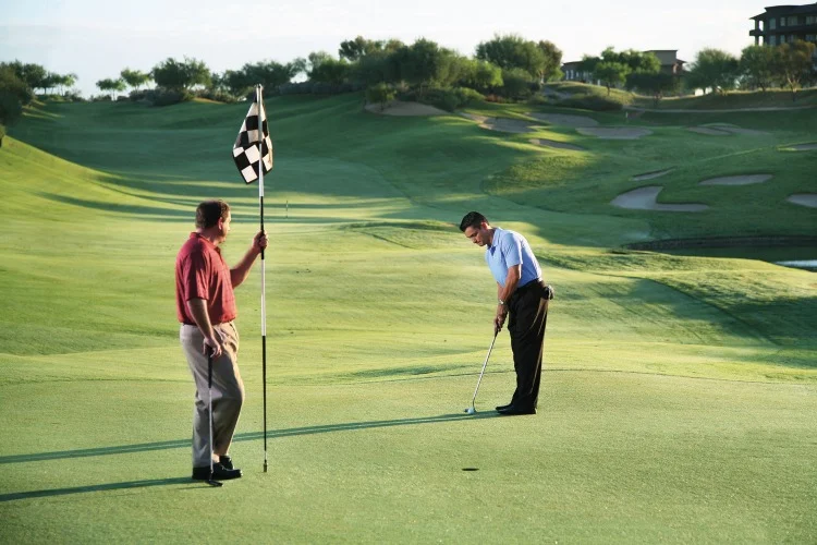 Twee mannen op een golfbaan waarvan de een de vlag vasthoudt terwijl de ander een bal richting de hole putt