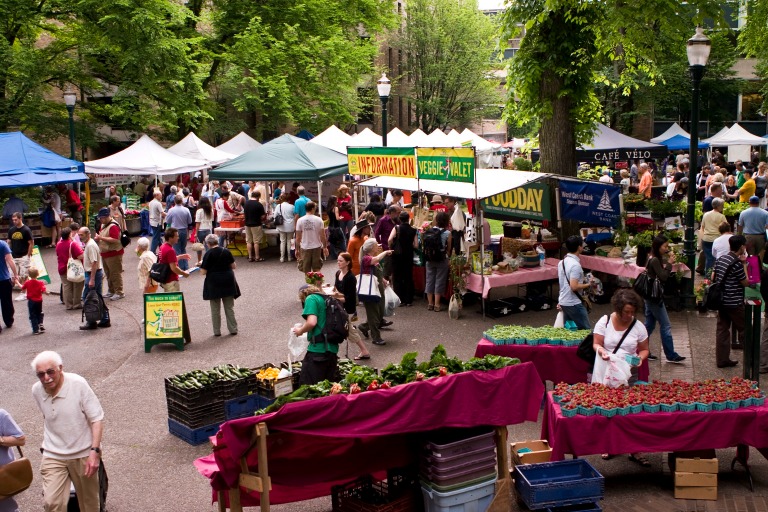 Drukke boerenmarkt in Portland met kraampjes vol groenten en fruit onder tenten en bomen