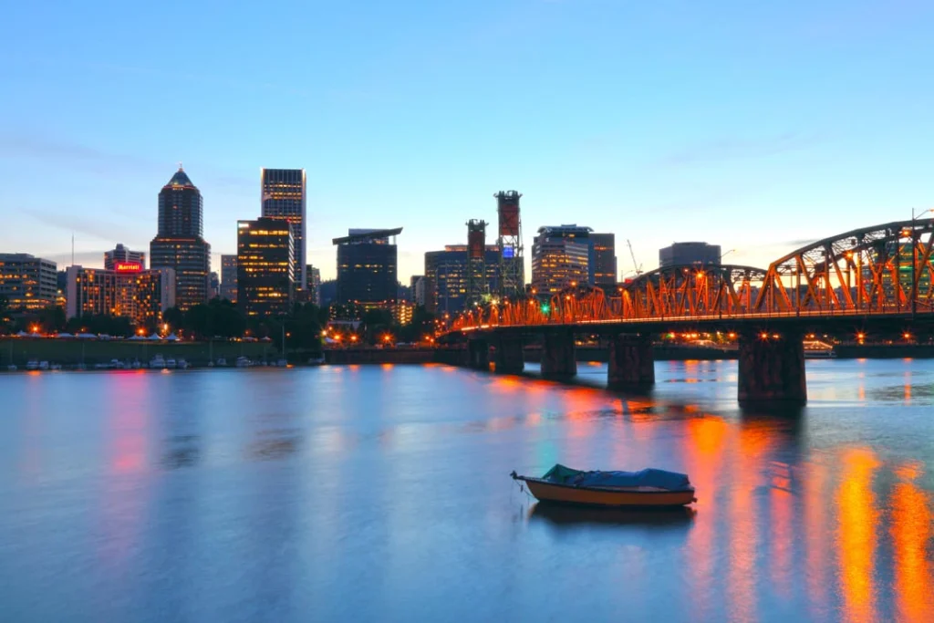 Verlichte wolkenkrabbers en een ijzeren brug weerspiegelen in het water van de Willamette rivier in Portland bij zonsondergang, met een klein bootje op de voorgrond