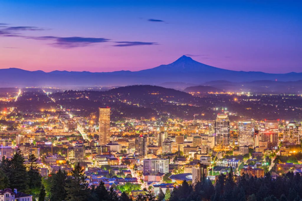 Verlichte gebouwen van Portland bij invallende schemering, met de silhouet van Mount Hood zichtbaar boven de heuvels