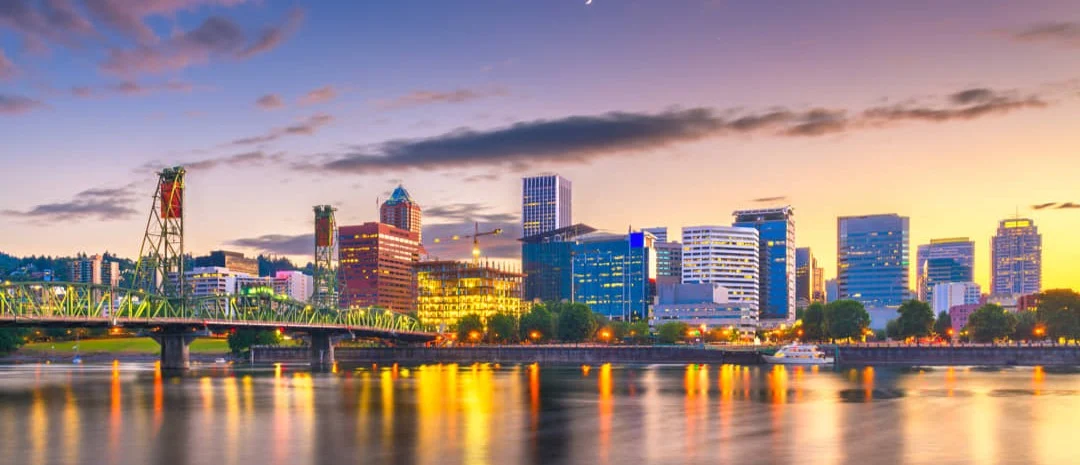 Skyline van een Amerikaanse stad met een verlichte ophaalbrug over een rivier terwijl de lucht roze en oranje kleurt bij zonsondergang