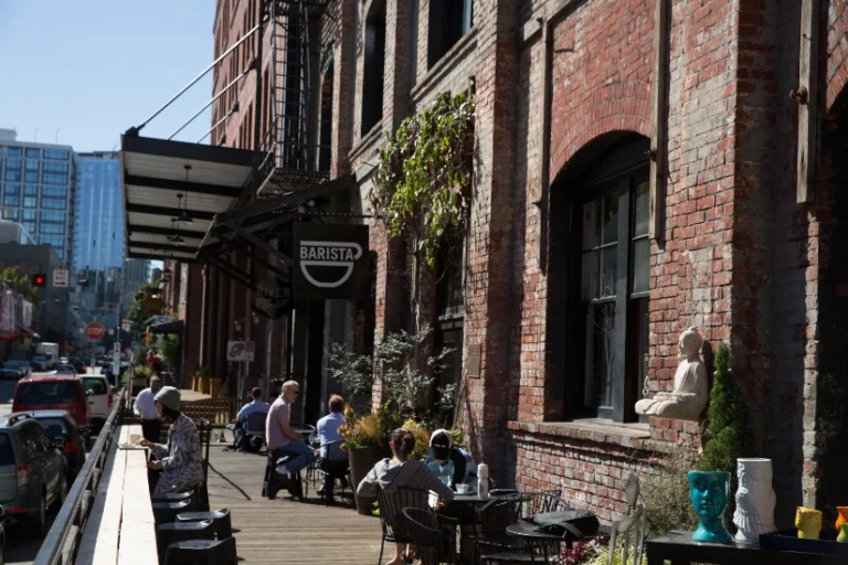 Mensen zitten op een zonnig terras met tafeltjes voor een bakstenen pakhuis met een Barista bord in de Pearl District van Portland
