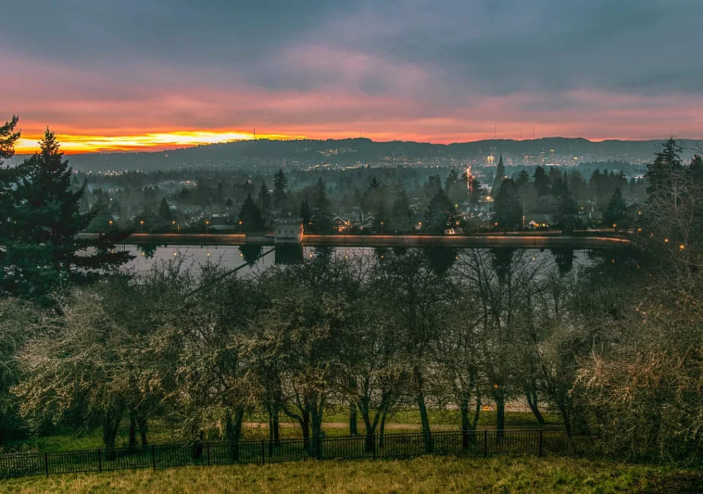 Kale bomen bij een hek op de voorgrond met uitzicht over een rivier en de verlichte skyline van Portland bij een gloeiende oranje zonsondergang