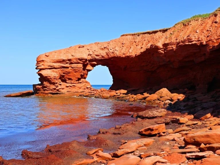 Natuurlijke boog van rode zandsteen aan de kustlijn met kalm blauw water en verspreide rotsblokken op het strand op Prince Edward Island
