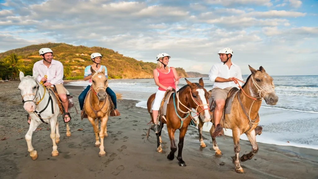 Vier ruiters met helm rijden op paarden langs de branding van een strand bij zonsondergang in Punta Islita, Guanacaste