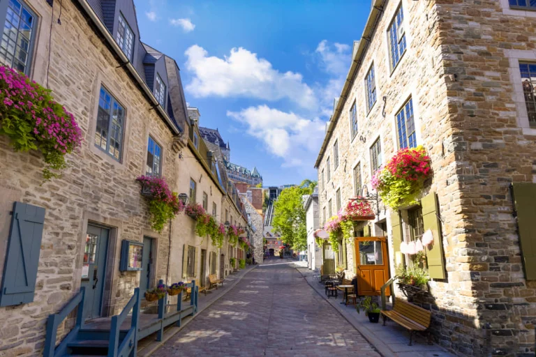 Stenen huizen met bloembakken vol roze bloemen langs een geplaveid straatje in de wijk Petit Champlain in Québec City