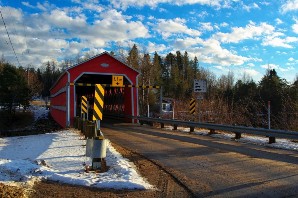 Traditionele rode houten overdekte brug met gele waarschuwingsstrepen staat aan een besneeuwde weg tegen een blauwe lucht