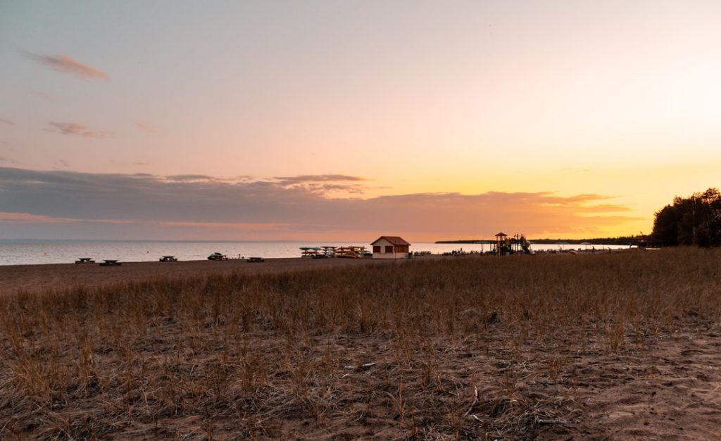 Zandstrand met droog helmgras op de voorgrond en een klein wit paviljoen bij een steiger tegen een oranje avondlucht