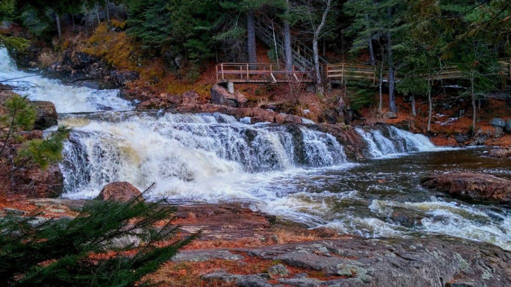 Brede waterval stort zich over rotsen terwijl bezoekers vanaf een houten platform tussen herfstbomen toekijken