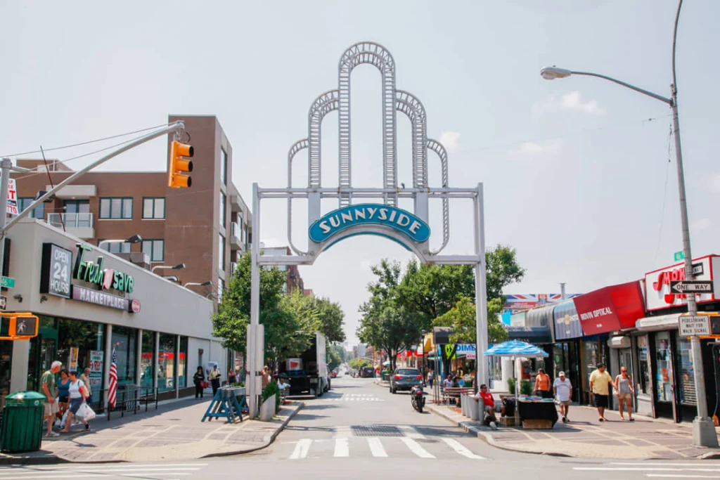Sierlijke metalen boog met het opschrift Sunnyside boven een winkelstraat in Queens met winkels en voetgangers