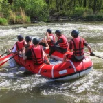Een groep mensen met helm en zwemvest peddelt in een rode rubberboot door kolkend rivierwater omgeven door groen