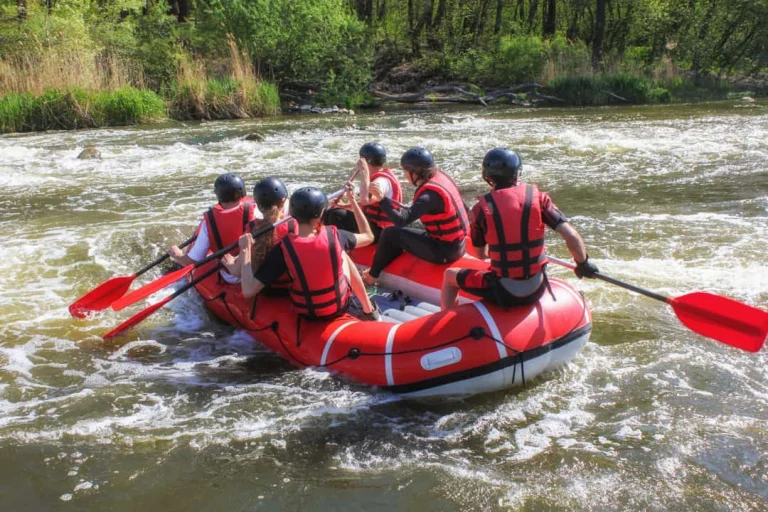 Een groep mensen met helm en zwemvest peddelt in een rode rubberboot door kolkend rivierwater omgeven door groen