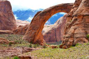 Enorme natuurlijke stenen boog spant over een canyonvloer, met bezoekers klein afgebeeld onderaan en wolken achter de rotsen