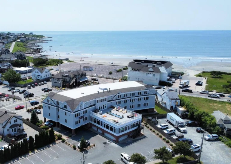Luchtfoto van een wit strandhotel met plat dak naast een parkeerplaats, met een lang zandstrand en de zee op de achtergrond in Rhode Island