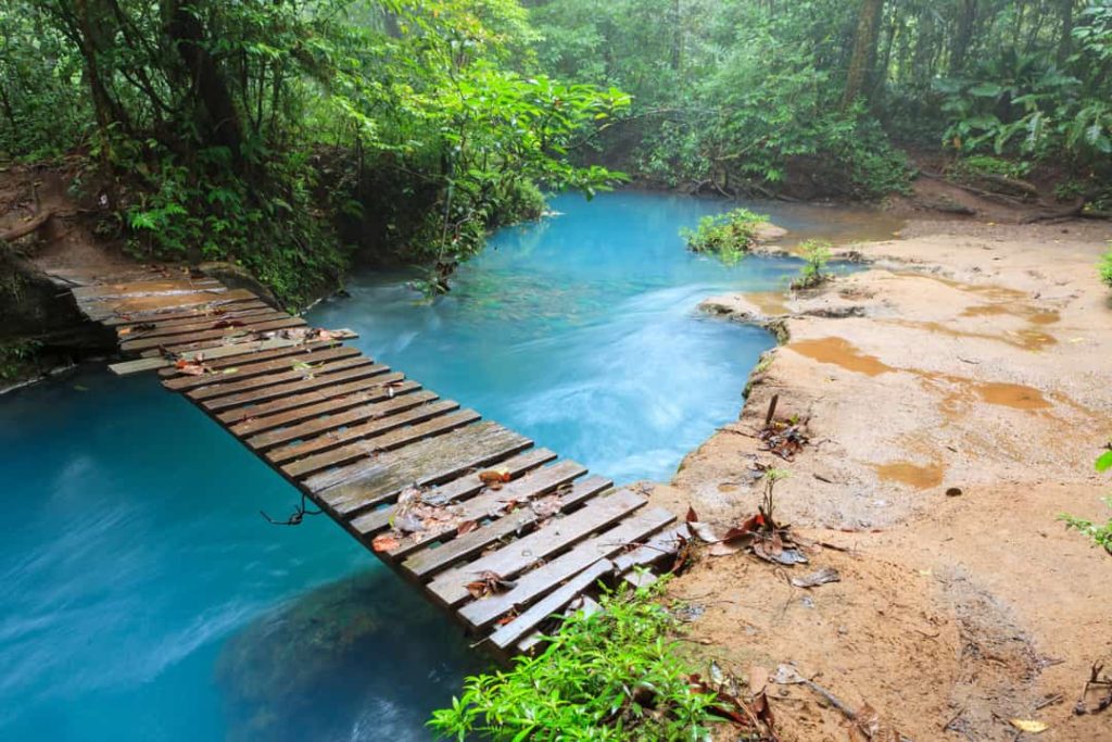 Smalle houten brug met leuning over een felturkooize beek, omringd door zanderige oevers en regenwoud bij Rio Celeste in Costa Rica
