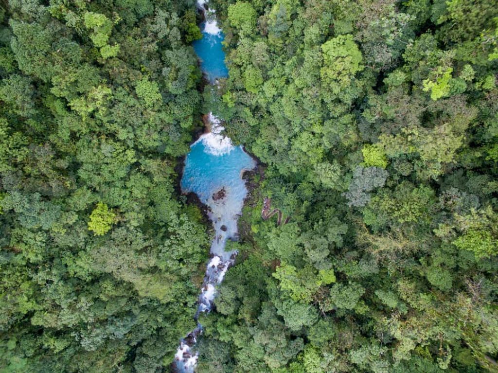 Luchtfoto recht van boven op een felturkooize poel en kleine waterval, omringd door dicht groen bladerdak bij Rio Celeste in Costa Rica