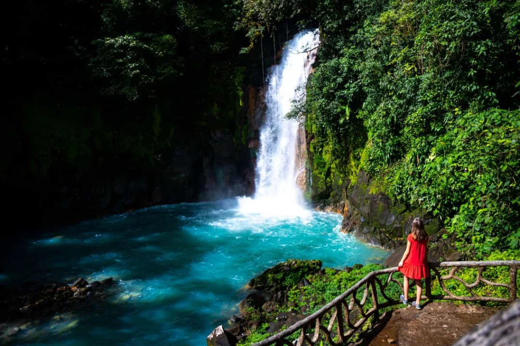 Vrouw in een rode jurk staat op een uitkijkpunt en kijkt naar de waterval die neerstort in een turkooize poel bij Rio Celeste in Costa Rica