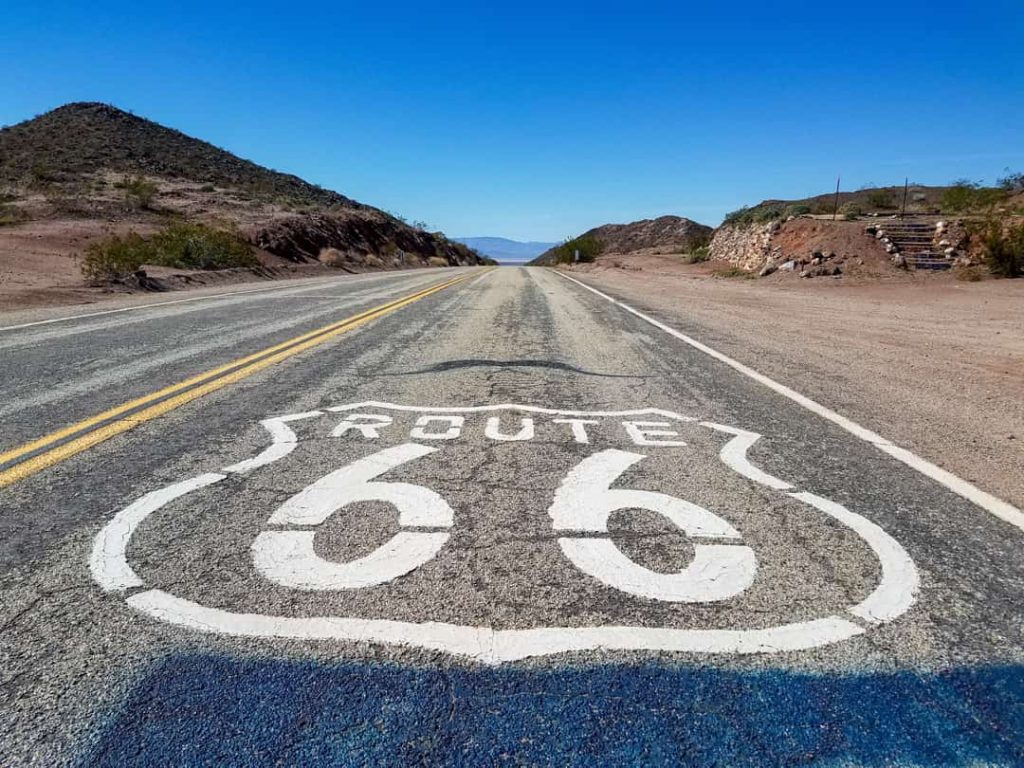 Wit geschilderde route 66 markering op het wegdek van een lege tweebaansweg tussen kale woestijnheuvels