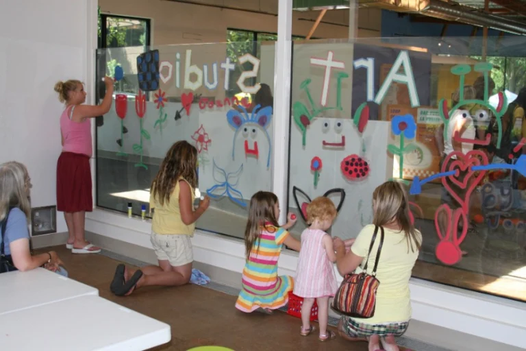 Kinderen en een begeleidster tekenen met verf op een groot raam met de tekst Art Studio in een kindermuseum