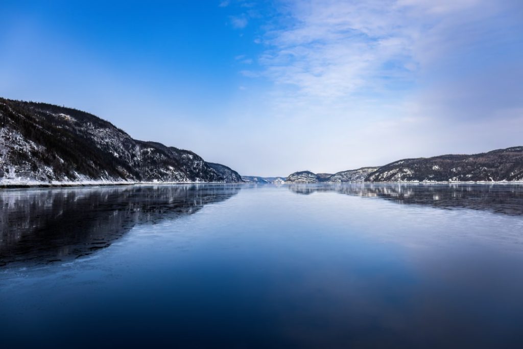 Bevroren fjord met besneeuwde oevers strekt zich uit onder een lichtblauwe winterlucht met wat sluierwolken