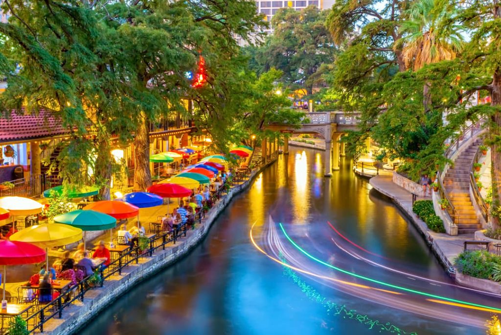 Verlichte terrassen met kleurrijke parasols langs het kanaal van de River Walk in San Antonio, met lichtsporen van varende boten