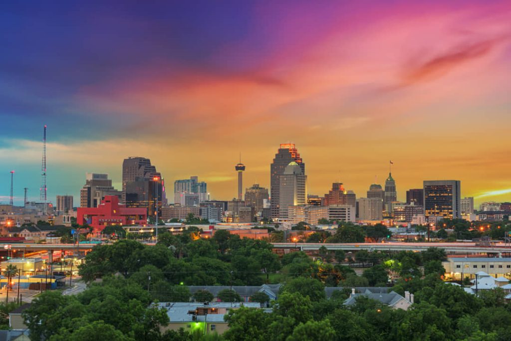 Skyline van het centrum van San Antonio met de Tower of the Americas onder een kleurrijke avondlucht, met bomen op de voorgrond