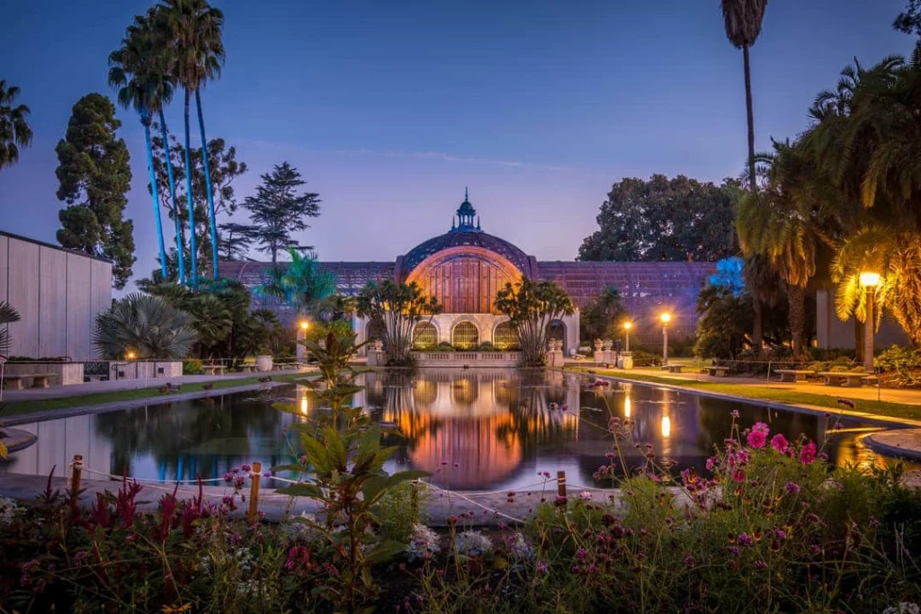 Verlicht koepelgebouw van glas en hout weerspiegelt in een vijver omringd door palmbomen en bloemen in Balboa Park in San Diego